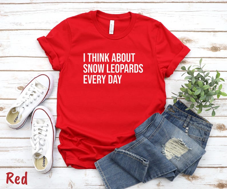May include: A red t-shirt with the text "I THINK ABOUT SNOW LEOPARDS EVERY DAY" in white. The shirt is paired with blue jeans and white sneakers. The word "Red" is at the bottom left.