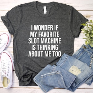 May include: Dark gray t-shirt with the text "I WONDER IF MY FAVORITE SLOT MACHINE IS THINKING ABOUT ME TOO" in white. The shirt is paired with blue jeans and white sneakers.