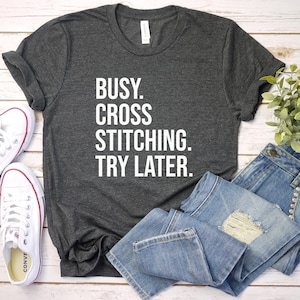 May include: A charcoal grey t-shirt with the words "BUSY. CROSS STITCHING. TRY LATER." in white. The image includes blue jeans, white trainers with red trim, and a small green plant. The items are arranged on a wooden surface.