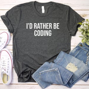 May include: A dark gray t-shirt with the text "I'd rather be coding" in white lettering. The shirt is laid out on a white wooden surface with a pair of white sneakers and a pair of blue jeans.