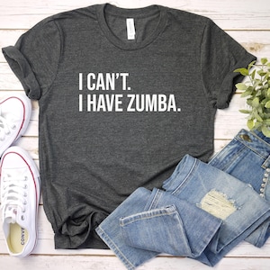 May include: Dark gray t-shirt with the text "I CAN'T. I HAVE ZUMBA." in white. The shirt is paired with blue jeans and white sneakers with red accents. A small green plant is also in the image.