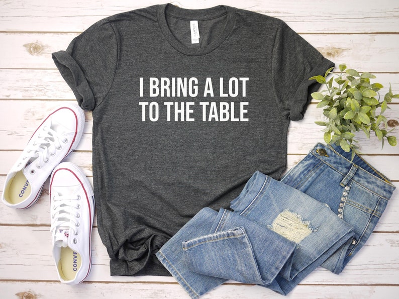Może przedstawiać: Szary t-shirt z nadrukiem "I bring a lot to the table" w kolorze białym na przodzie. Koszulka leży płasko na białym drewnianym tle z parą białych but&oacute;w sportowych i niebieskimi dżinsami.