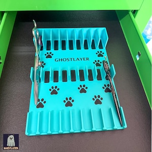 Op de afbeelding: Een turquoise organizer voor verzorgingsgereedschap met de tekst "GHOSTLAYER" en pootafdrukken. Twee verzorgingsgereedschappen zijn in de organizer geplaatst. De organizer staat in een groene lade.