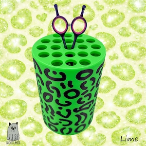 Puede incluir: Un organizador cilíndrico verde lima con diseño de estampado de leopardo negro, que sostiene un par de tijeras moradas y rosas. El organizador tiene múltiples agujeros en la parte superior. La palabra "Lime" está en la esquina inferior derecha.
