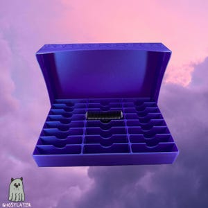 Könnte beinhalten: Eine lila Aufbewahrungsbox mit offenem Deckel, die mehrere Fächer zeigt. Ein schwarzes Objekt liegt in der Mitte. Die Box befindet sich vor einem bewölkten, lila-rosa Himmel.