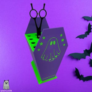 Könnte beinhalten: Ein lila-grüner Sarg-Organizer mit Geister- und Stern-Design, der eine schwarze Schere hält. Der Organizer trägt den Text "GHOSTLAYER" auf der Vorderseite. Schwarze Fledermaus-Ausschnitte befinden sich im Hintergrund.
