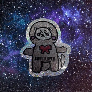 Könnte beinhalten: Ein silberner Aufkleber mit einem Cartoon-Bären in einem Raumanzug mit roter Fliege. Der Aufkleber hat den Text "GHOSTLAYER" unten. Der Hintergrund ist eine Galaxie aus blauen, lila und weißen Sternen.