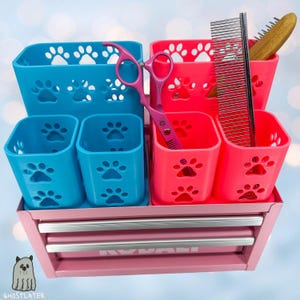 Könnte beinhalten: Eine rosa Metall-Werkzeugkiste enthält blaue und pinkfarbene Behälter mit Pfotenabdruck-Dekor. Die Behälter enthalten Pflegeutensilien, darunter eine rosa Schere und Kämme. Das Ghostlayer-Logo befindet sich unten links.