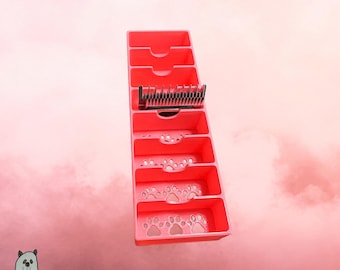 May include: A pink plastic organizer tray featuring multiple rectangular compartments with cutout paw print designs on the base. One compartment holds a black metal clipper guard attachment with multiple teeth. The organizer is vertical.