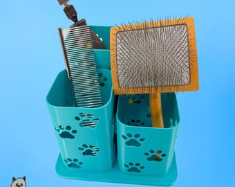 Op de afbeelding: Een blauwgroene plastic organizer voor dierverzorgingshulpmiddelen met uitgesneden pootafdrukpatronen. De organizer bevat een metalen kam, een roterende nagelslijper en een rechthoekige houten slickerborstel met fijne metalen haren. Tekst GHOSTLAYER.