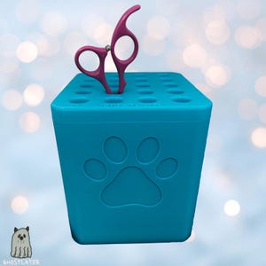 May include: A teal grooming tool organizer with a paw print design and a pair of pink grooming shears. The organizer has multiple holes for storing tools. The shears are made of metal and have a curved handle.