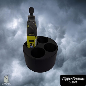 May include: A black cylindrical tool holder featuring four circular openings, with one yellow and grey rotary tool inserted into a slot. The base is solid black, and the text "Clipper/Dremel Insert" is displayed in a black box at the bottom right.