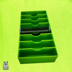 Könnte beinhalten: Ein leuchtend grüner Organizer mit mehreren Fächern, der einen silbernen Kamm enthält. Der Organizer hat eine rechteckige Form mit geschwungenen Aussparungen. Der Kamm befindet sich in der Mitte. Der Hintergrund ist einfarbig lindgrün.