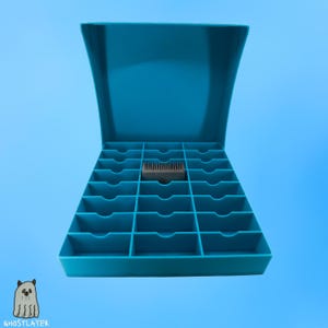 Könnte beinhalten: Eine türkisblaue Organizer-Box mit einem Klappdeckel, der geöffnet ist, um mehrere Fächer freizulegen. Ein kleines, graues Objekt liegt in der Mitte. Die Box befindet sich vor einem hellblauen Hintergrund. Das Ghostlayer-Logo befindet sich unten links.