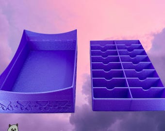 Op de afbeelding: Een paarse opbergset bestaande uit een grote open bak met een pootafdrukpatroon aan de voorzijde en een apart rechthoekig inzetstuk met zestien kleine vakjes voorzien van gebogen uitsparingen voor gemakkelijke toegang.