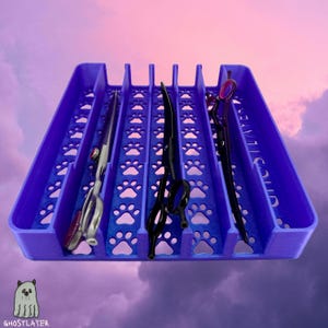 Könnte beinhalten: Ein lila Organizer für Tierpflege-Werkzeuge mit Pfotenabdruck-Ausschnitten, der drei Scherenpaare für die Fellpflege enthält. Die Scheren sind schwarz und silberfarben, mit roten und violetten Akzenten. Der Organizer hat den Text "GHOSTLAYER".