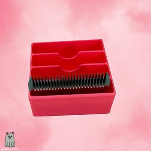 Könnte beinhalten: Ein leuchtend roter Organizer mit drei Fächern und einem Schlitz für eine schwarz-silberne Clipperklinge. Der Organizer besteht aus Kunststoff und befindet sich vor einem rosa Hintergrund. Das Produkt ist für Pflege-Werkzeuge konzipiert.