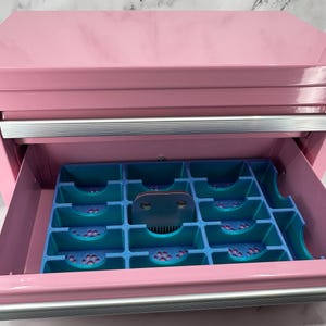 Op de afbeelding: Een roze metalen opbergkist met een uitschuifbare lade met een blauwe en groenblauwe 3D-geprinte inzetbak. De inzetbak heeft meerdere rechthoekige vakken met uitsparingen in de vorm van pootafdrukken op de bodem en een metalen scheerkop.