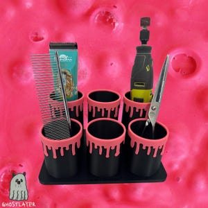 Könnte beinhalten: Ein schwarzer Organizer mit sechs zylindrischen Fächern, jedes mit einem rosa Tropf-Farben-Design. Er enthält Pflegeutensilien, darunter ein Kamm, ein Clipper, eine Schere und ein Rotationswerkzeug. Der Hintergrund ist eine helle, strukturierte rosa Oberfläche.