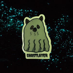 Könnte beinhalten: Ein gestanzter Aufkleber mit einer cartoonartigen, geisterhaften Kreatur mit bärenartigen Ohren und einer braunen Nase und Augen. Der Aufkleber trägt unten den Text "GHOSTLAYER". Der Hintergrund ist schwarz mit leuchtenden blauen Punkten.