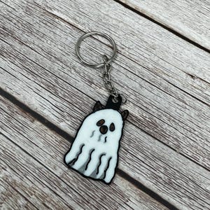 Könnte beinhalten: Ein weißer Geister-Schlüsselanhänger mit schwarzen Augen und einem schwarzen Mund. Der Geist trägt einen schwarzen Hut und Hörner. Der Schlüsselanhänger ist an einem silbernen Schlüsselring befestigt.