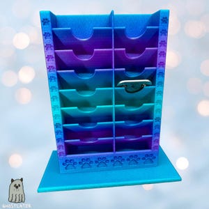 Könnte beinhalten: Ein mehrfarbiger Schreibtisch-Organizer mit einem Farbverlauf aus Blau, Lila und Türkis. Der Organizer hat mehrere Schlitze und ist mit Pfotenabdrücken verziert. Ein kleiner, silberfarbener Gegenstand befindet sich in einem der Schlitze. Die Basis ist einfarbig blau.