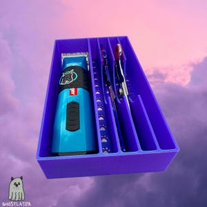 Könnte beinhalten: Ein lila Organizer mit Fächern für Pflegeutensilien. Ein blau-schwarzer Haarschneider für Haustiere mit einem Hunde-Motiv befindet sich im ersten Fach. Silberne Pflegeschere und andere Werkzeuge befinden sich in den anderen Fächern. Der Organizer hat Pfotenabdruck-Ausschnitte.