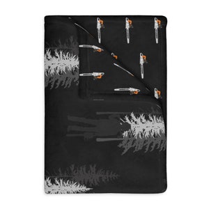 May include: Black fleece blanket with a repeating pattern of gray pine trees and orange and white chainsaws. The blanket is folded in half, revealing the same pattern on the other side.