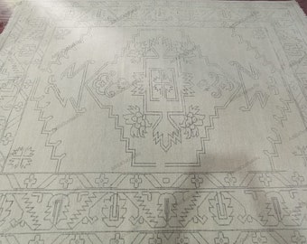 May include: A light beige rug with a geometric pattern in a darker shade of beige. The pattern features a central cross shape with floral motifs and a border of repeating geometric shapes.