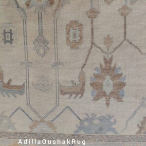 Op de afbeelding: Een beige Oushak-kleed met een traditioneel bloemenpatroon in tinten bruin en blauw. Het kleed heeft een franje rand.