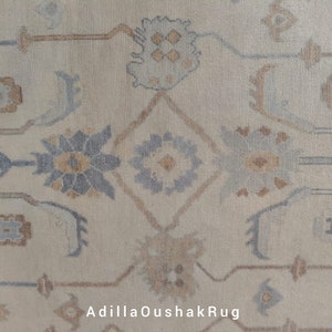 Op de afbeelding: Close-up van een beige tapijt met een traditioneel Perzisch ontwerp. Het tapijt heeft ingewikkelde patronen in tinten blauw, bruin en beige. De tekst "AdiilaOushakRug" is zichtbaar in de rechterbenedenhoek.