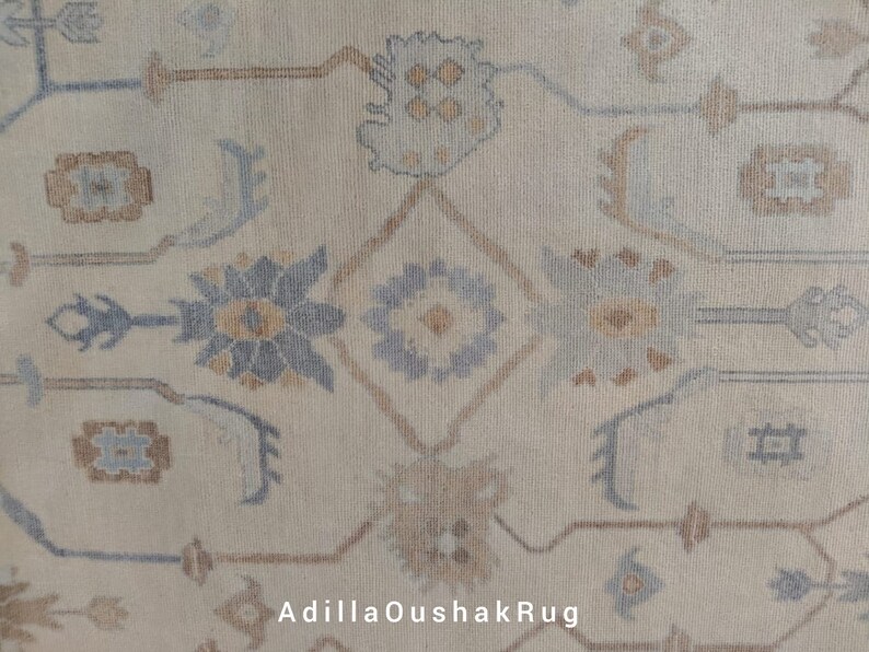 Op de afbeelding: Close-up van een beige tapijt met een traditioneel Perzisch ontwerp. Het tapijt heeft ingewikkelde patronen in tinten blauw, bruin en beige. De tekst "AdiilaOushakRug" is zichtbaar in de rechterbenedenhoek.