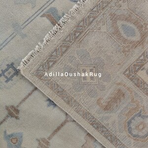 Op de afbeelding: Een close-up van een beige en blauw gepatroneerd tapijt met een franje rand. Het tapijt heeft een subtiel geometrisch ontwerp en is gemaakt van een zacht, geweven materiaal. De tekst "AdiilaOushakRug" is zichtbaar in de afbeelding.