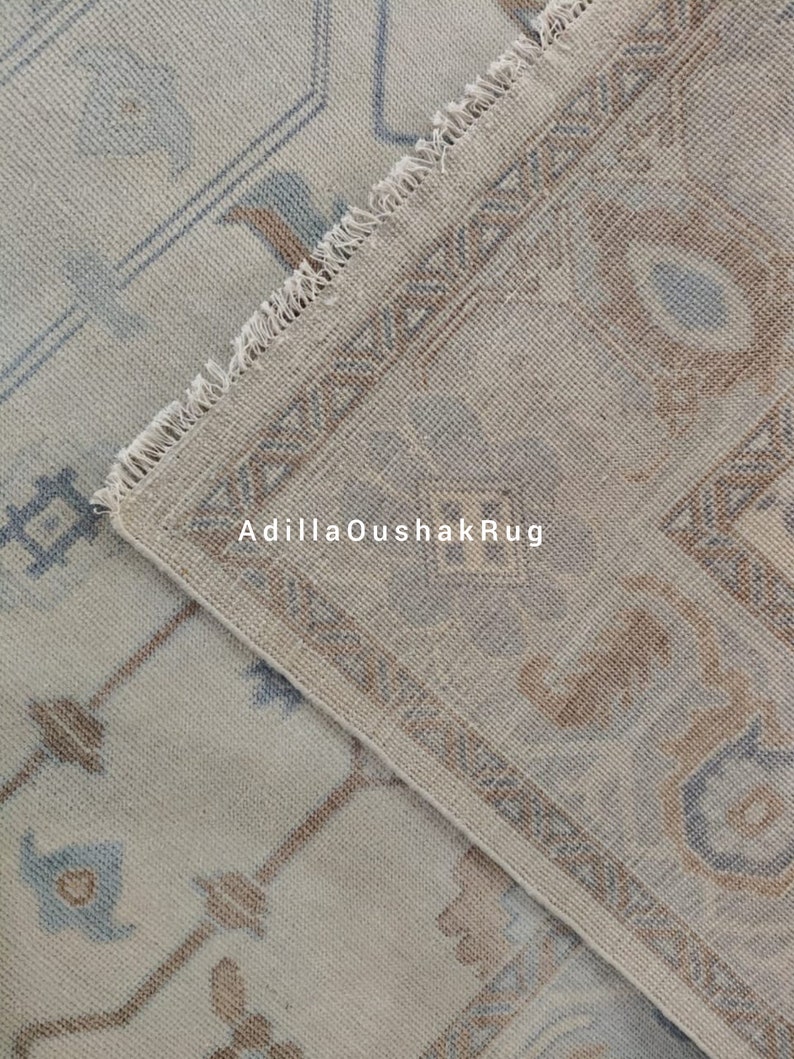 Op de afbeelding: Een close-up van een beige en blauw gepatroneerd tapijt met een franje rand. Het tapijt heeft een subtiel geometrisch ontwerp en is gemaakt van een zacht, geweven materiaal. De tekst "AdiilaOushakRug" is zichtbaar in de afbeelding.