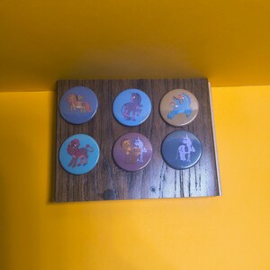 Op de afbeelding: Zes ronde buttons gerangschikt in twee rijen van drie. Elke button is voorzien van een unieke cartoon pony-illustratie in diverse kleuren, waaronder paars, blauw, bruin en beige, op een rechthoekig displaybord met een donkere houtnerftextuur.