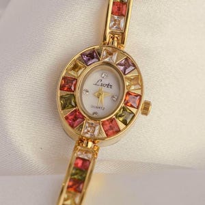 May include: A gold-toned quartz watch with a white face and colorful square gemstones. The watch features the text "Lvotr" and "QUARTZ" on the face. The band is also adorned with colorful gemstones.