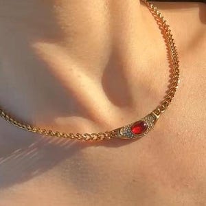 May include: A gold-toned chain necklace featuring a central crescent-shaped pendant. The pendant is set with a smooth, oval-shaped red stone at its center, surrounded by small, clear, sparkling stones embedded along the curved metal frame.