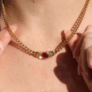 May include: A gold-toned chain necklace featuring a central horizontal bar element. The centerpiece is adorned with a smooth, oval-shaped deep red gemstone flanked by small, clear, sparkling stones set into the metallic surface of the bar.