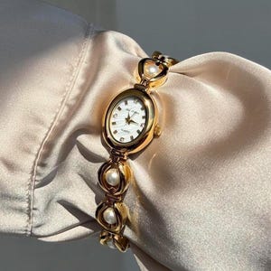 May include: Gold-toned wristwatch with an oval face and a white dial. The watch features a gold bracelet with pearl accents. The watch is displayed on a cream-colored fabric.