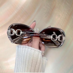 Puede incluir: Gafas de sol ovaladas con lentes tintadas marrones y detalles met&aacute;licos plateados en las patillas. La montura es de color marr&oacute;n transl&uacute;cido. Las gafas de sol est&aacute;n sujetas por una persona.