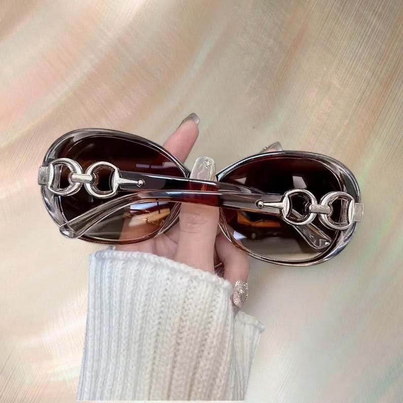 Puede incluir: Gafas de sol ovaladas con lentes tintadas marrones y detalles met&aacute;licos plateados en las patillas. La montura es de color marr&oacute;n transl&uacute;cido. Las gafas de sol est&aacute;n sujetas por una persona.