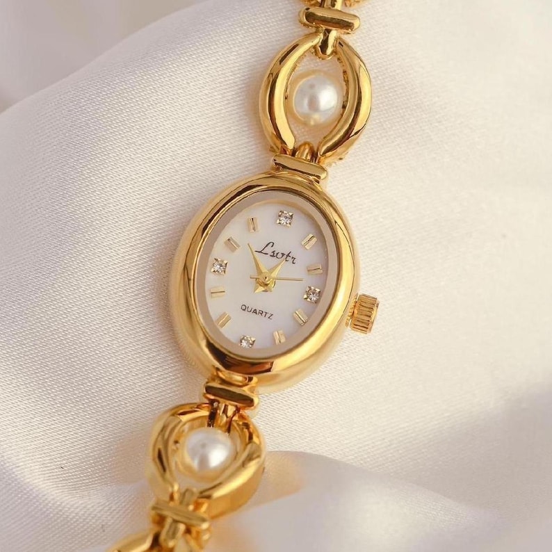 May include: Gold-toned quartz watch with an oval face and a white dial. The watch features the text "Liotr" and "Quartz". The watch band has decorative links with embedded pearls. The watch is set against a white background.