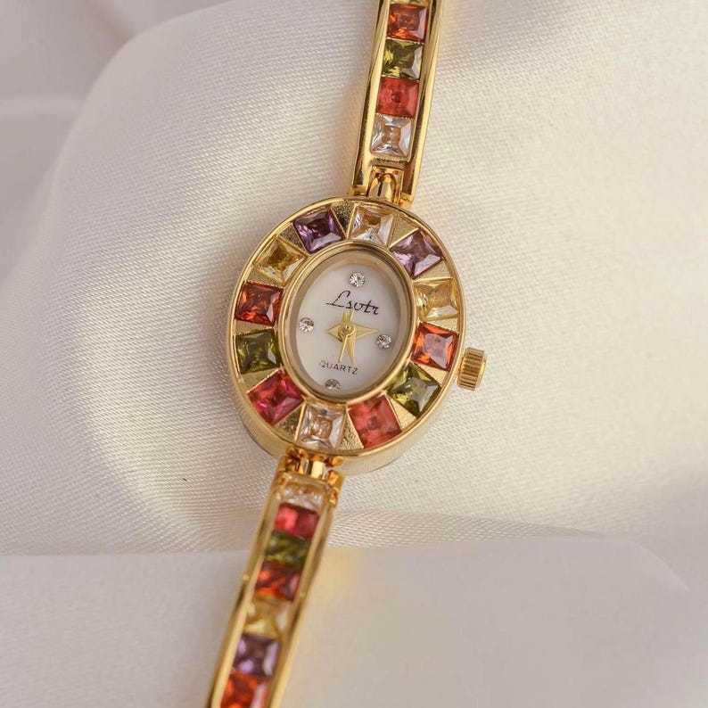 May include: A gold-toned quartz watch with a white face and colorful square gemstones. The watch features the text "Lvotr" and "QUARTZ" on the face. The band is also adorned with colorful gemstones.