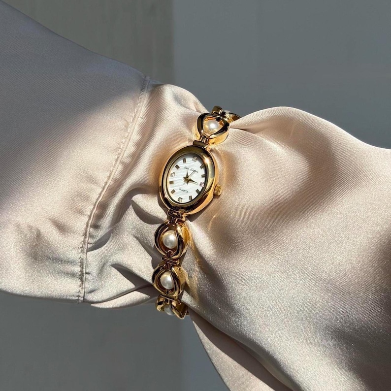 May include: Gold-toned wristwatch with an oval face and a white dial. The watch features a gold bracelet with pearl accents. The watch is displayed on a cream-colored fabric.