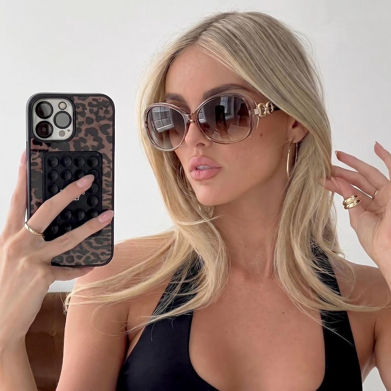 Puede incluir: Gafas de sol marrones de gran tama&ntilde;o con detalles dorados en las patillas. Las lentes tienen un tinte degradado. La persona sostiene un tel&eacute;fono con una funda con estampado de leopardo y un agarre emergente negro. La persona lleva pendientes de aro dorados y un top negro.