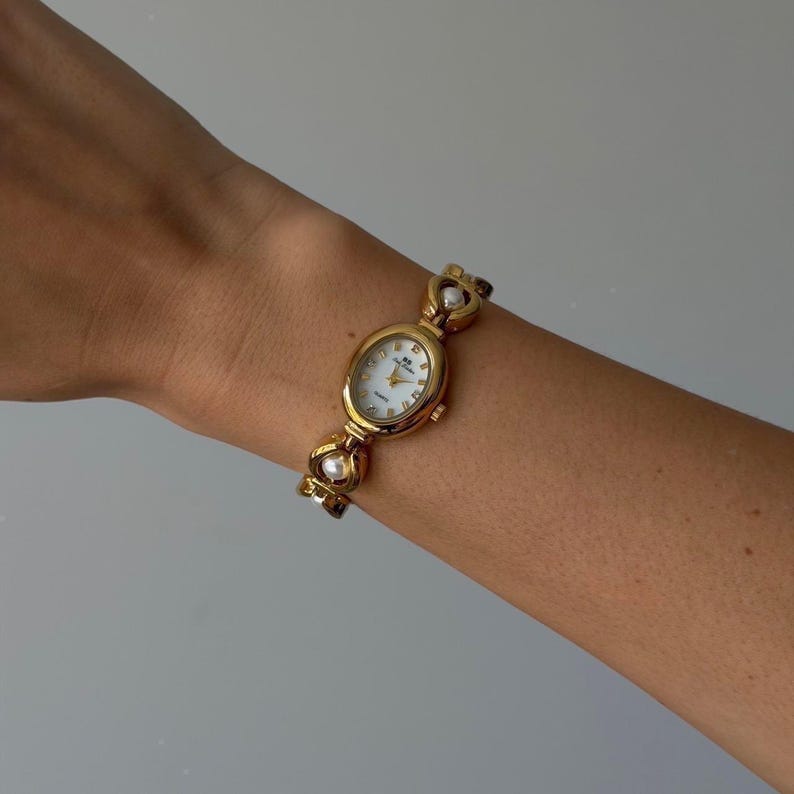 May include: Gold-toned wristwatch with an oval face and a white dial. The watch features a gold band with heart-shaped links and faux pearl accents. The watch is displayed on a wrist against a neutral background.