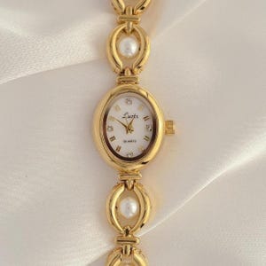 May include: Gold-toned quartz watch with an oval face and a linked bracelet. The watch face is white with gold hands and Roman numerals. The bracelet features pearl accents. The watch has the word "Quartz" printed on the watch face.