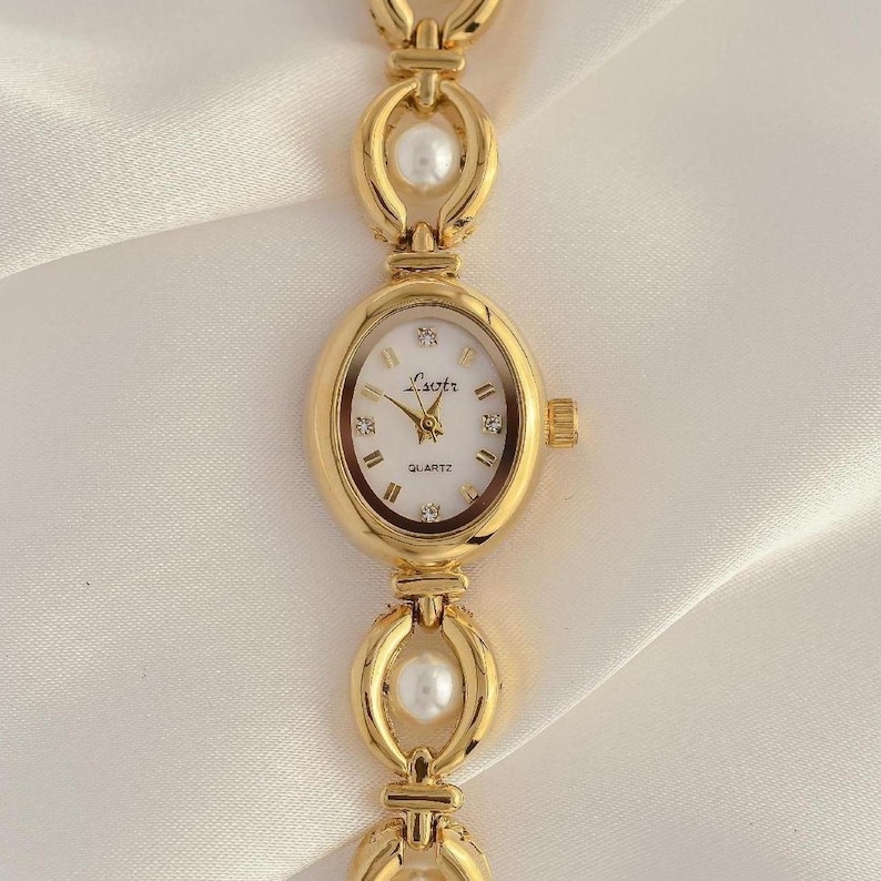 May include: Gold-toned quartz watch with an oval face and a linked bracelet. The watch face is white with gold hands and Roman numerals. The bracelet features pearl accents. The watch has the word "Quartz" printed on the watch face.