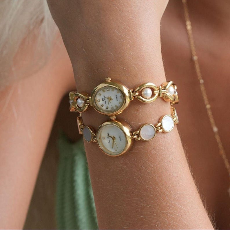 May include: Two gold-toned wristwatches with oval faces and white dials. The top watch features a pearl-accented band, while the bottom watch has a band with iridescent, circular accents. Both watches are on a wrist.