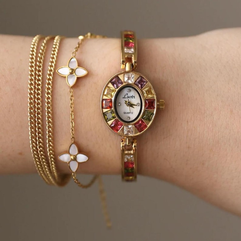 May include: A gold-tone quartz watch with a white face labeled Lsotr, surrounded by a bezel of multicolored square-cut stones. The watch is paired with a gold-tone chain bracelet featuring two white four-petaled flower charms and a curb chain.